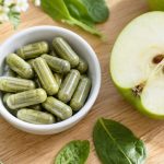 Capsule verzi de supliment într-un bol alb, lângă o jumătate de măr verde