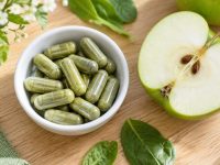 Capsule verzi de supliment într-un bol alb, lângă o jumătate de măr verde